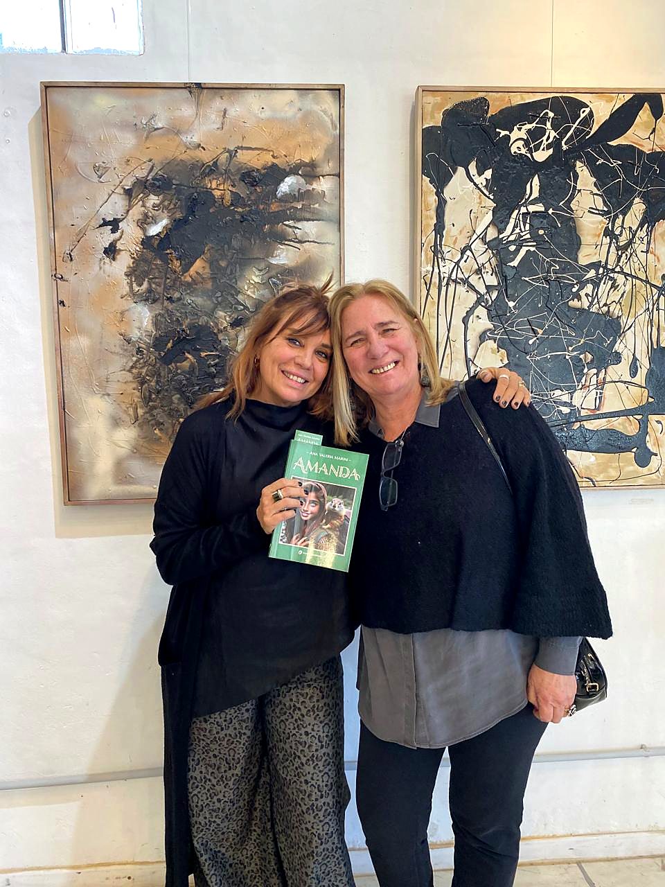 Vale Marini y una visitante sostienen el libro Amanda frente a dos pinturas matéricas enmarcadas