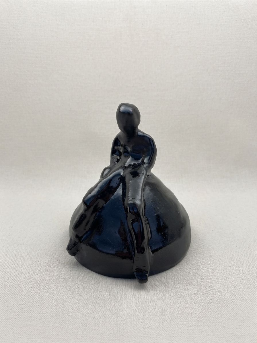 Figura de cerámica negra sentada al borde de una loma redondeada con las piernas colgando