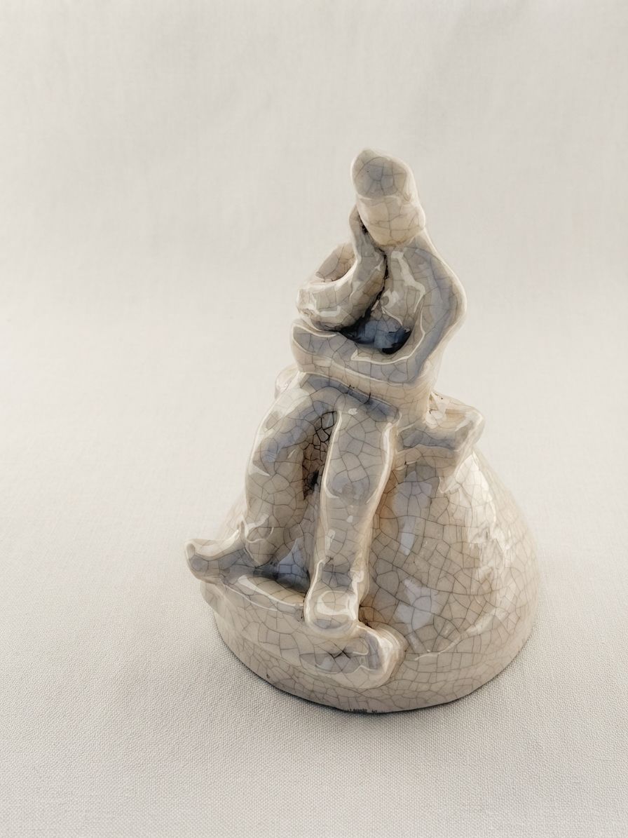 Pequeña figura de cerámica clara con esmalte craquelado, sentada sobre una colina, con la mano en el mentón